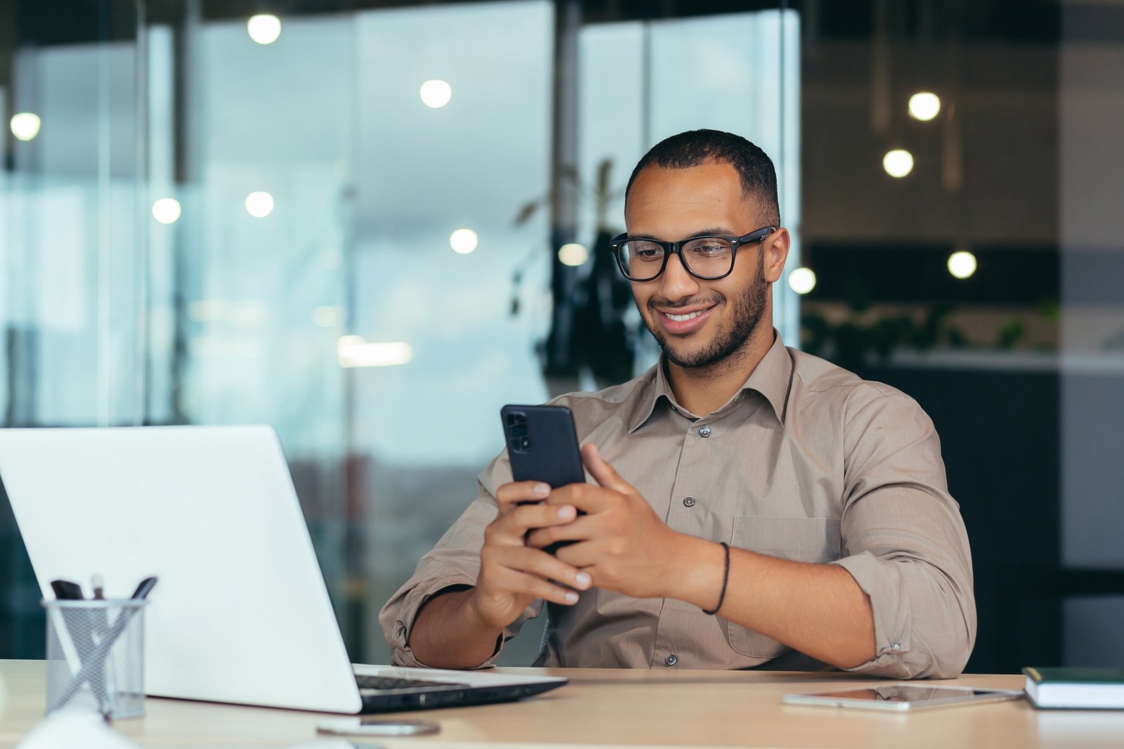 Um empresário afro-americano feliz, de óculos, trabalhando em um escritório moderno com um laptop.